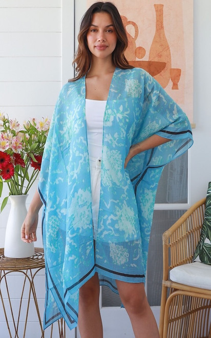 Model wearing the Lagoon Ripple Kimono. The lightweight, semi-sheer sky blue kimono features an abstract ripple print with a thin dark navy border, styled casually over a solid white top and white shorts.