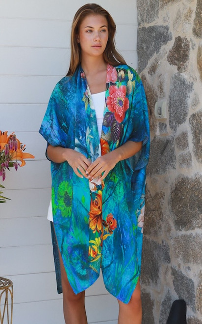 Model wearing the Amazonian Reef kimono. The lightweight, semi-sheer kimono features a vibrant turquoise and deep blue background with bright orange and coral floral print, styled casually over a solid white top.