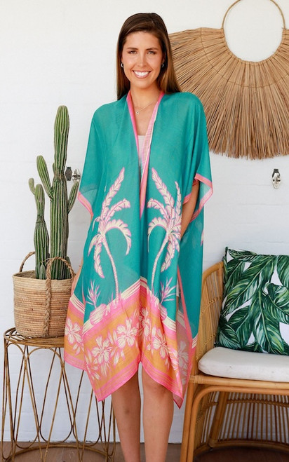 Model wearing the easy, open-front Malibu Sunset kimono. The vibrant teal chiffon kimono features a bold pattern of pink and cream palm trees with an orange and pink hibiscus border along the hem, styled over a casual neutral top.