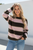 Model is wearing a cosy wool blend striped knit in pink and dark tones with a relaxed fit, paired with light blue jeans outdoors