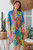 wearing the easy, open-front Botanical Oasis kimono. The vibrant cornflower-blue chiffon cardigan features a bold pattern of large yellow, green, and pink tropical flowers, styled over a neutral tank top.