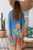 wearing the easy, open-front Botanical Oasis kimono. The vibrant cornflower-blue chiffon cardigan features a bold pattern of large yellow, green, and pink tropical flowers, styled over a neutral tank top.