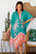 Model wearing the easy, open-front Malibu Sunset kimono. The vibrant teal chiffon kimono features a bold pattern of pink and cream palm trees with an orange and pink hibiscus border along the hem, styled over a casual neutral top.