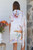 Model wearing the easy, open-front Sahara Bloom kimono. The white lightweight fabric features an abstract pattern of muted lavender, sage green, and ochre yellow leaves, finished with a detailed periwinkle and orange geometric border along the hem, styled casually over a solid white tank and white shorts.