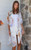 Model wearing the easy, open-front Sahara Bloom kimono. The white lightweight fabric features an abstract pattern of muted lavender, sage green, and ochre yellow leaves, finished with a detailed periwinkle and orange geometric border along the hem, styled casually over a solid white tank and white shorts.