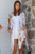 Model wearing the easy, open-front Sahara Bloom kimono. The white lightweight fabric features an abstract pattern of muted lavender, sage green, and ochre yellow leaves, finished with a detailed periwinkle and orange geometric border along the hem, styled casually over a solid white tank and white shorts.