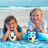 Little kids smiling while relaxing with the Bluey and Bingo Pillow Pets at the beach.