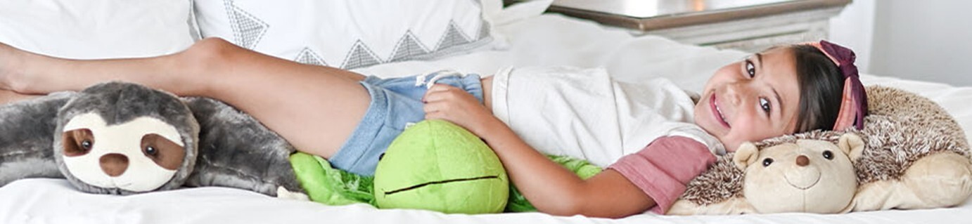 Little girl laying on her bed all comfy cozy and smiling with three Pillow Pets.