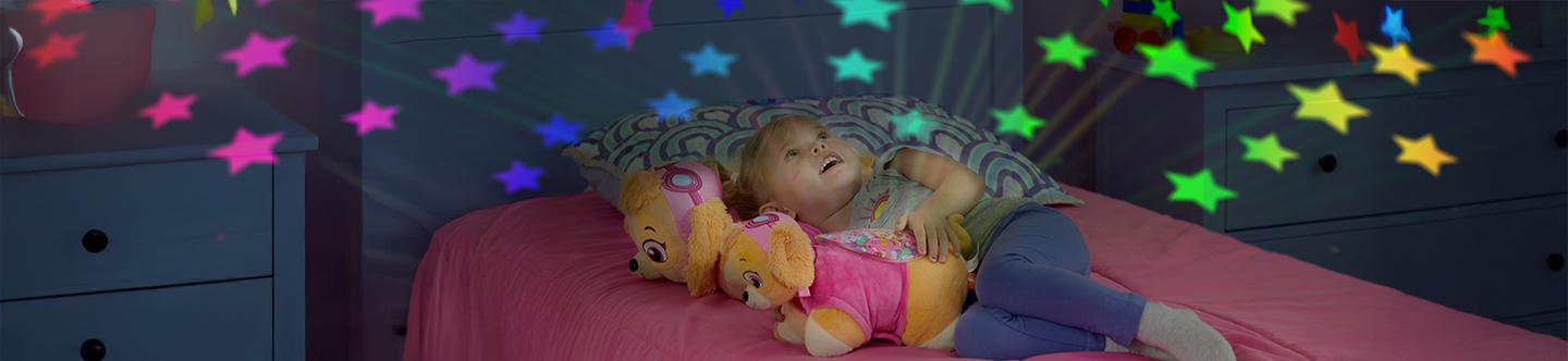A little girl with a Skye Sleeptime Lite on her bed, with her room all lit up with colorful rainbow stars.