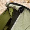 Snugpak Scorpion 3 IX olive tent deployed on gravel against stone ruins with door fly open showing front porch storage area with backpack inside and rolled mosquito net visible at top of door opening
