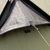 Snugpak Scorpion 3 IX olive tent pitched on gravel against stone ruins showing full fly-closed exterior profile with opposing pole structure, Snugpak branding on flysheet body, and guy lines staked out
