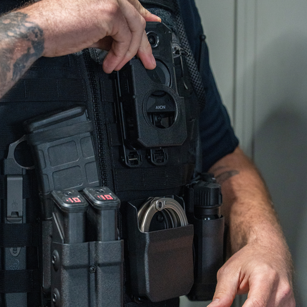 Close-up of a hand reaching for the Zero9 body cam case mounted on a plate carrier with mag pouches