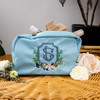 Custom embroidered cosmetic bag personalized with the bride's wedding crest. Wedding crest embroidery is done in smokey blue, navy, green and pink threads. Cosmetic bag is shown on a woven table with flowers and cosmetic items for decoration.