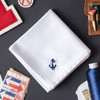 White handkerchief embroidered with a small anchor in navy thread in the corner. Handkerchief shown on gray background with stamps and thread.