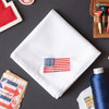 White handkerchief embroidered with a small American flag in red, white and blue in the corner. Handkerchief shown on gray background with stamps and thread.
