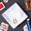 White handkerchief embroidered with a small horseshoe in matte gold in the corner. Handkerchief shown on gray background with stamps and thread.