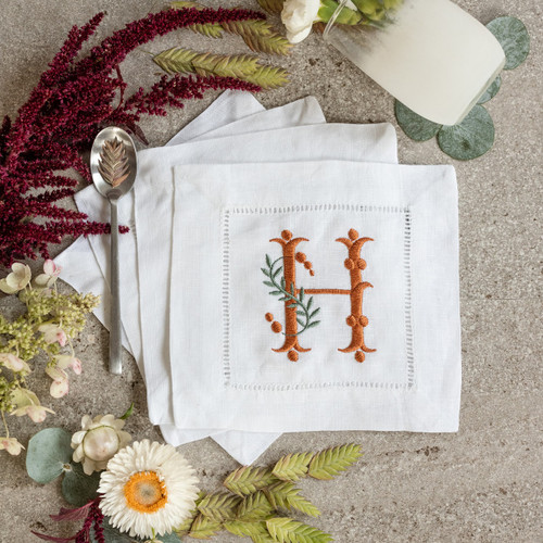 Custom embroidered cocktail napkins. Set of 4. Customized with a monogram initial in burnt orange thread and sage colored leaves. Background has flowers, a glass and a spoon.