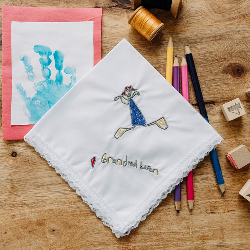 Embroidered kid's drawing on a white lace handkerchief. Embroidery is sewn in blue, black, yellow and red thread. Shown with color pencils, stamps, threads and kid's handprint painting for decoration.