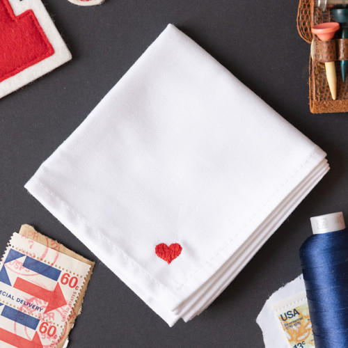 White handkerchief embroidered with a small red heart in the corner. Handkerchief shown on gray background with stamps, thread and golf tees.
