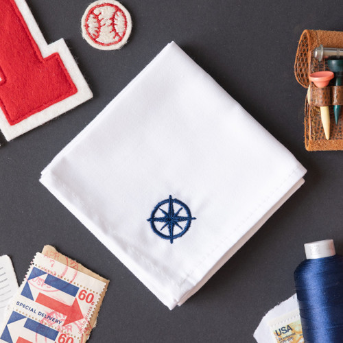 White handkerchief embroidered with a small elephant in navy in the corner. Handkerchief shown on gray background with stamps and thread.