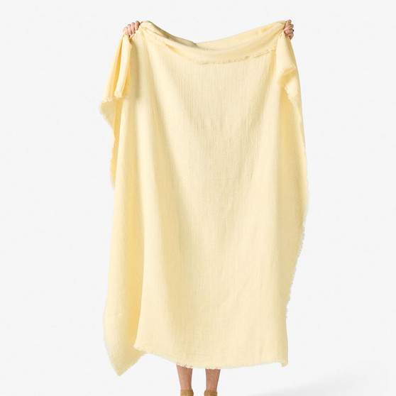 Linen House Maya Throw in Butter held up to show its full 130x170cm size, lightweight linen-cotton blend, and frayed edges.