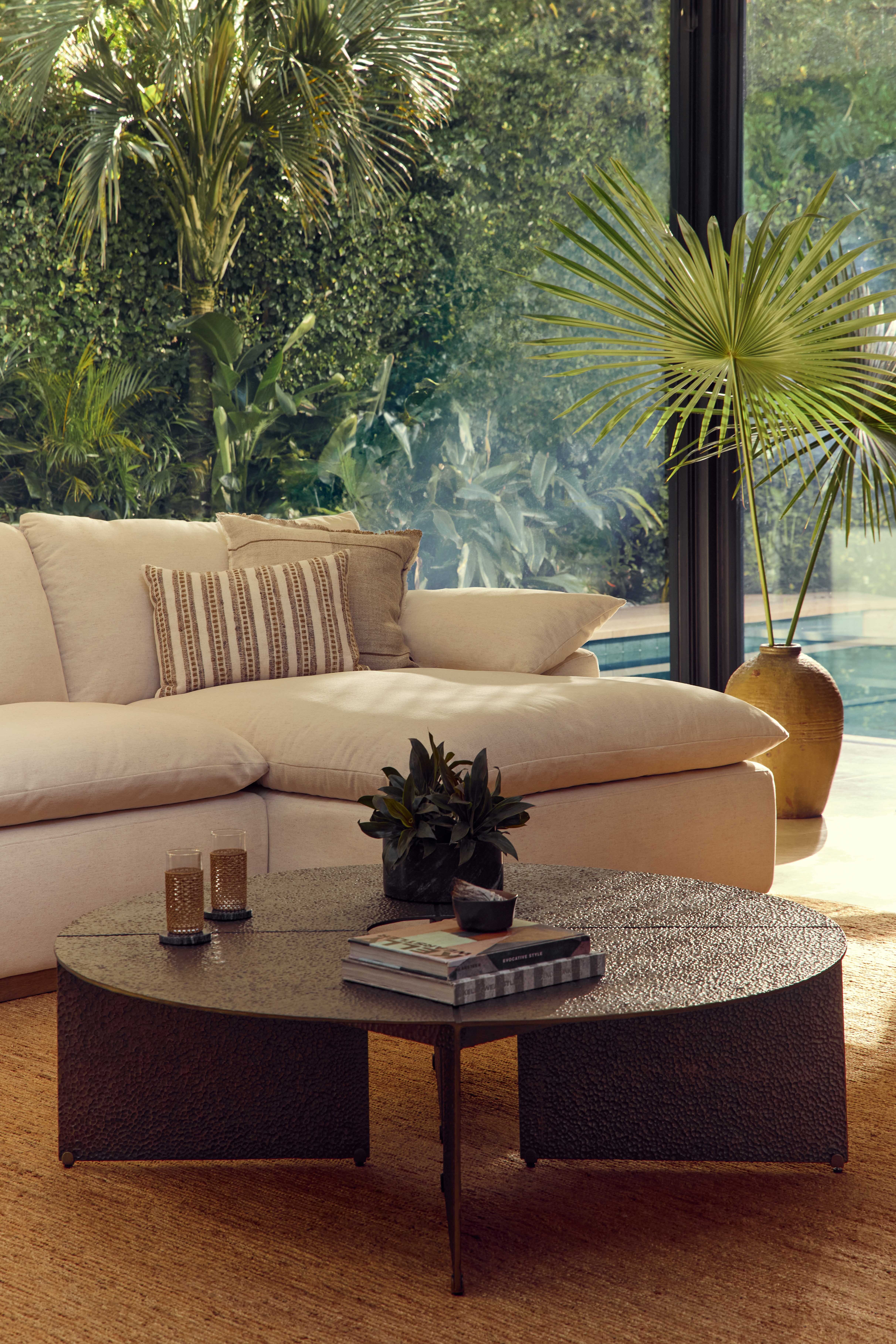Coco Republic Hermosa Modular Sofa in cream upholstery, styled beside a pool with lush greenery, woven cushions and a textured round coffee table.