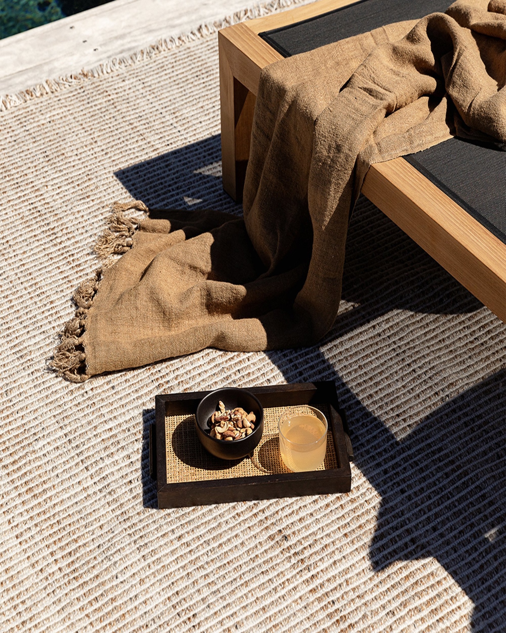  Outdoor rug in neutral weave, styled beside a teak sun lounger with throw and tray.