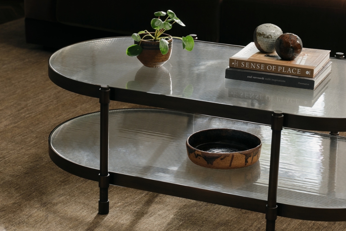 Oval coffee table with fluted glass shelves and bronze frame, styled with potted plant, decorative bowls, and stacked design books in a modern living room.