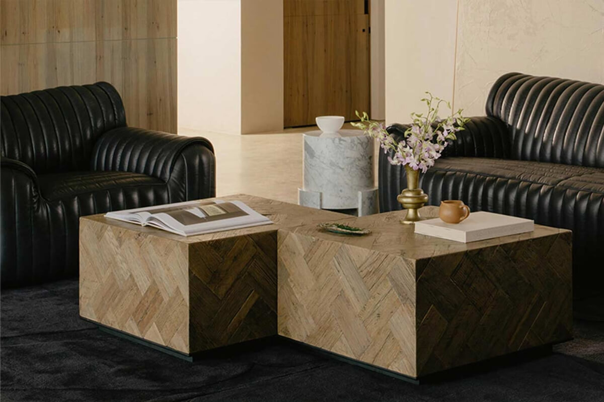 Square parquet timber coffee table with herringbone pattern, styled with books and a brass vase, set between black leather sofas in a contemporary living room.