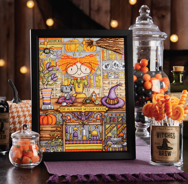 Project image from October of the Keepsake Calendar. Framed cross-stitched design of a  adorable witch in her shop selling potions, brooms, etc.