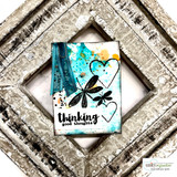 Mixed media card with the black dragonflies and hearts stamped over turquoise and orange ink splatters, a teal ribbon on a paper clip and a bold black sentiment at the bottom