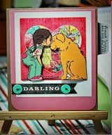 Bright card with a pink base and patterned paper heart, the girl coloured in a green top kissing a golden dog, with a darling label and a "first love" tab on the side.