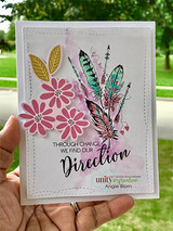 Card with the feathers in teal and gold beside stamped pink flowers and the through change we find our direction sentiment, held up outdoors.