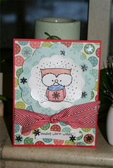 Card with the owl coloured in soft blue holding its mug inside a white cloud shape, over patterned circles with a red striped ribbon, sentiment "sending warm wishes."