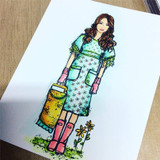 Card with Natalie watercolored in an aqua dress, pink gloves and pink rain boots with a yellow basket, standing on green grass beside small flowers.