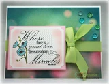 Great Love and Miracles card with the poppies colored in soft turquoise on a pink matted panel, set against a teal and pink bokeh background with a green ribbon and bow across the front.
