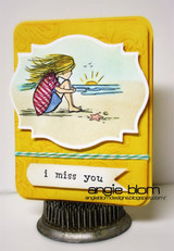 Yellow embossed card with a hand colored girl in a striped top sitting on the sand watching the sunrise, stamped 'i miss you' on a white banner.