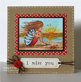 Kraft card with a red polka dot frame around a colored sunrise beach scene of a girl hugging her knees, stamped 'i miss you' on a tag with a red button.