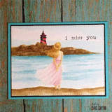 Watercolored card of a girl in a pink dress on the shore looking toward a lighthouse, stamped 'i miss you'.