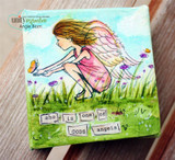 A canvas panel with the winged girl colored in pink and gold among wildflowers, with small stamped word tiles beneath her.