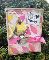 Mixed-media card photographed against greenery, the songbird in yellow and grey on a patterned panel with a pink ribbon and a 'Do your thing' tag.