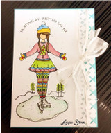 Card with Charlee colored in a blue and yellow hat, pink sweater, lime skirt and rainbow leggings on an icy pond, with a white ribbon and aqua cross-stitch border.