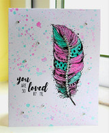 White splattered card with a feather coloured in magenta, teal and grey, tiny ink spatters around it and the sentiment "you ARE SO loved BY ME" stamped below.