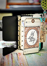 Journal page with 'It's the Little Things' stamped inside an ornate oval on a kraft tag tucked into a decorated album.