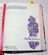 Bible page with a column of purple-blue flowers and 'We love because He first loved us'