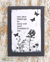 A wildflower silhouette rises across a script background inked in black, framed in grey — 'may your blessings be many and your troubles be few.'