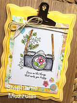 Card with the floral camera clipped to a yellow scalloped frame on wood.