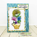 Yellow, pink and blue flowers in a glittery teacup on a white panel over blue patterned paper, stamped 'all we need is love...and coffee'.