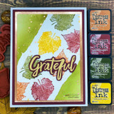 Green and blue watercolored panel stamped with leaf prints in yellow, coral and olive, gold embossed Grateful across the front, four Distress ink pads lined up beside the card.