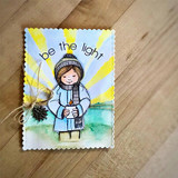 Card with the girl in soft blue against a sunrise of yellow rays on a scalloped panel with twine and a pinecone, and be the light at the top.